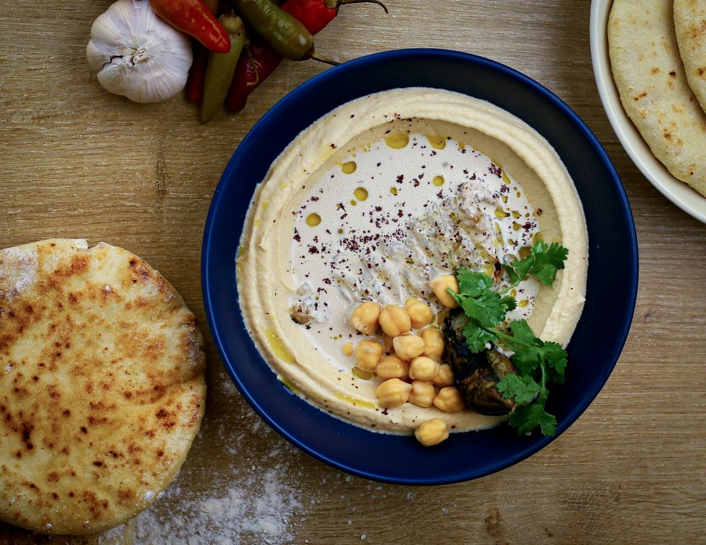 Délicieux houmous crémeux servi avec pois chiches entiers, aubergine grillée, coriandre fraîche et pain pita, sur une table en bois rustique. Une touche orientale gourmande et authentique.